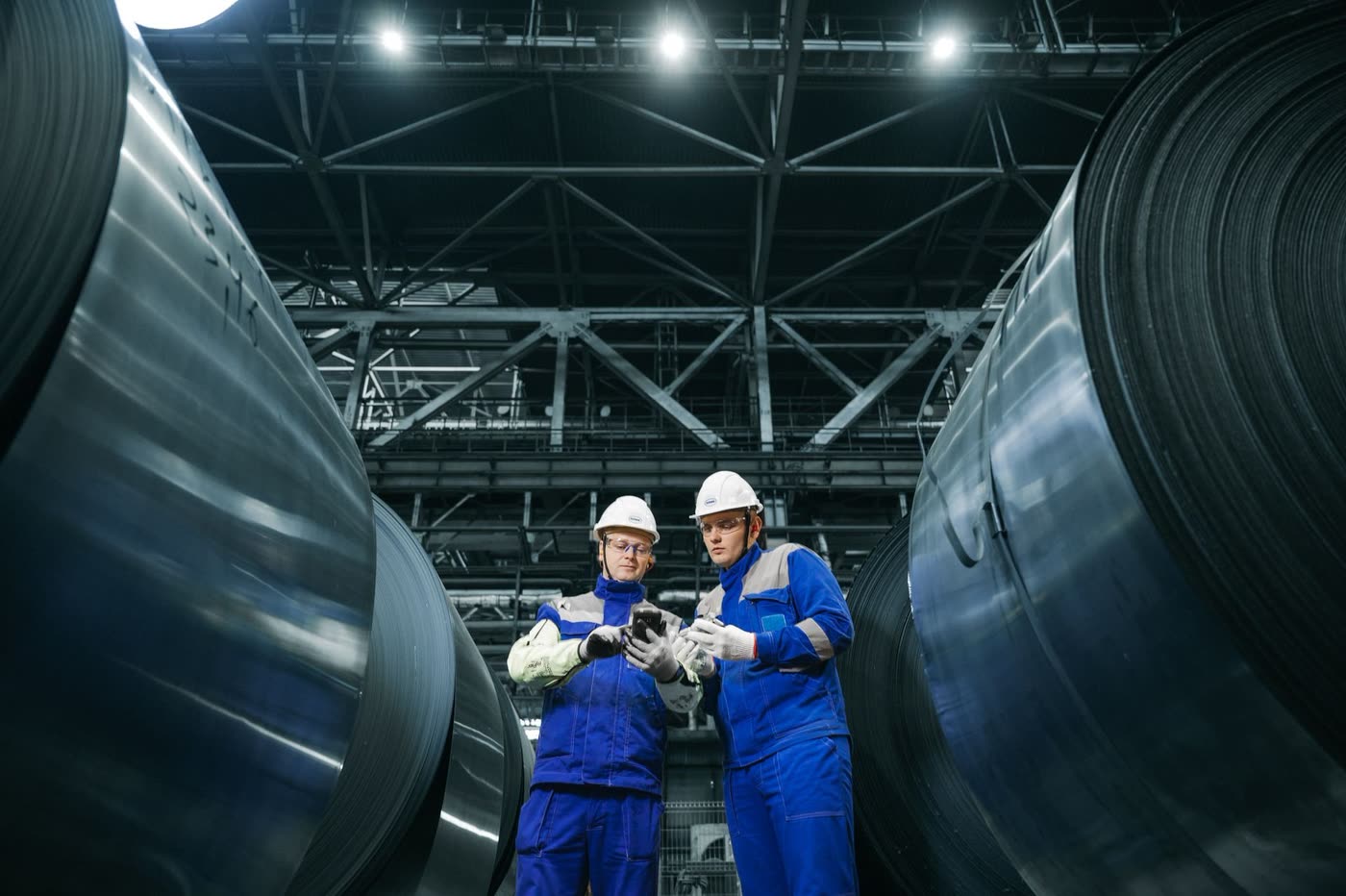Two engineers in work uniforms consulting over a handheld device between steel coils in a factory hall